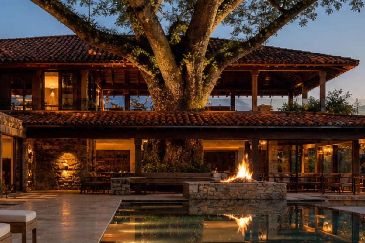 Hacienda al atardecer con árbol monumental atravesando el pabellón y fogón encendido junto a la piscina reflectante, registro de hospitalidad silenciosa